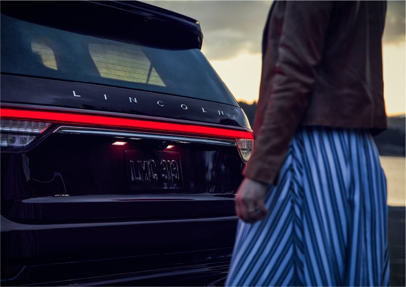 A person is shown near the rear of a 2023 Lincoln Aviator® SUV as the Lincoln Embrace illuminates the rear lights | All Star Lincoln in Prairieville LA